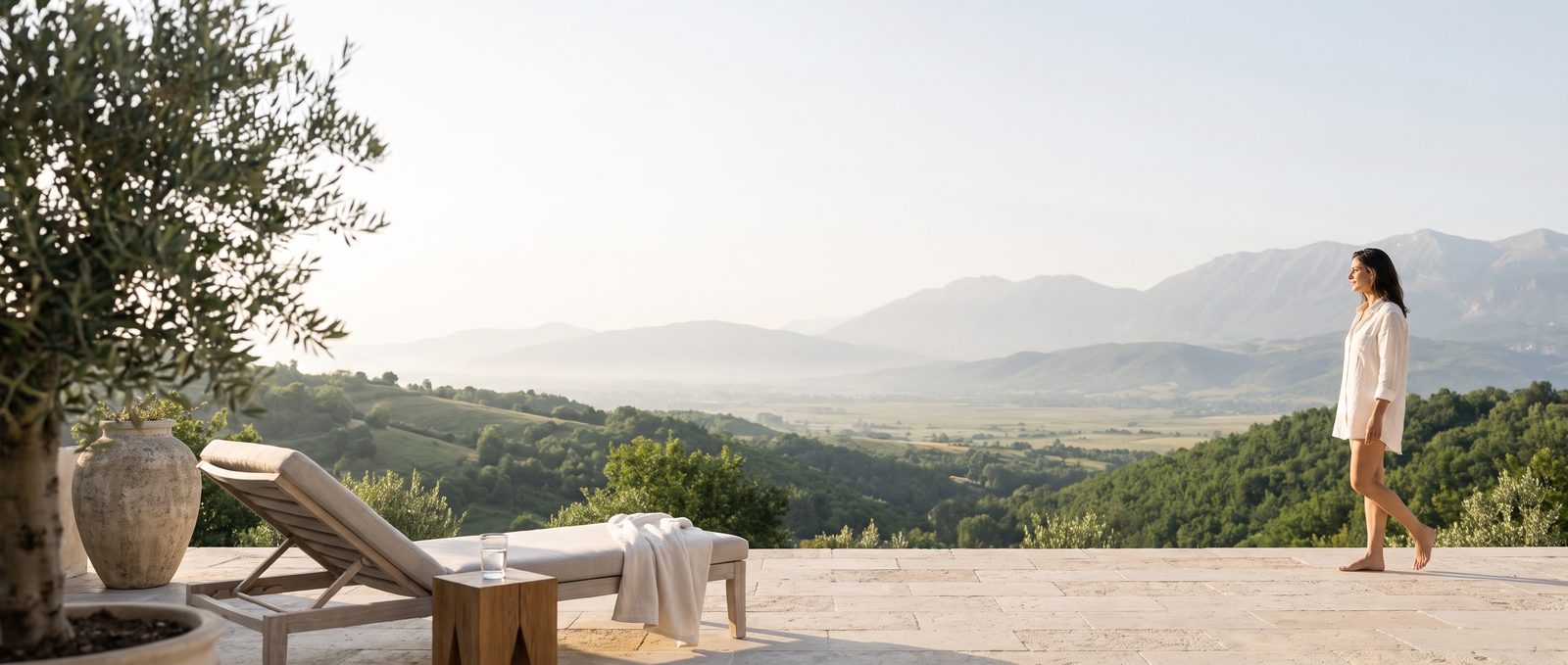 Eine ruhige Hotelterrasse am Morgen mit Blick auf grüne Hügel und Berge. Eine Frau im Leinenhemd geht auf die Aussicht zu.